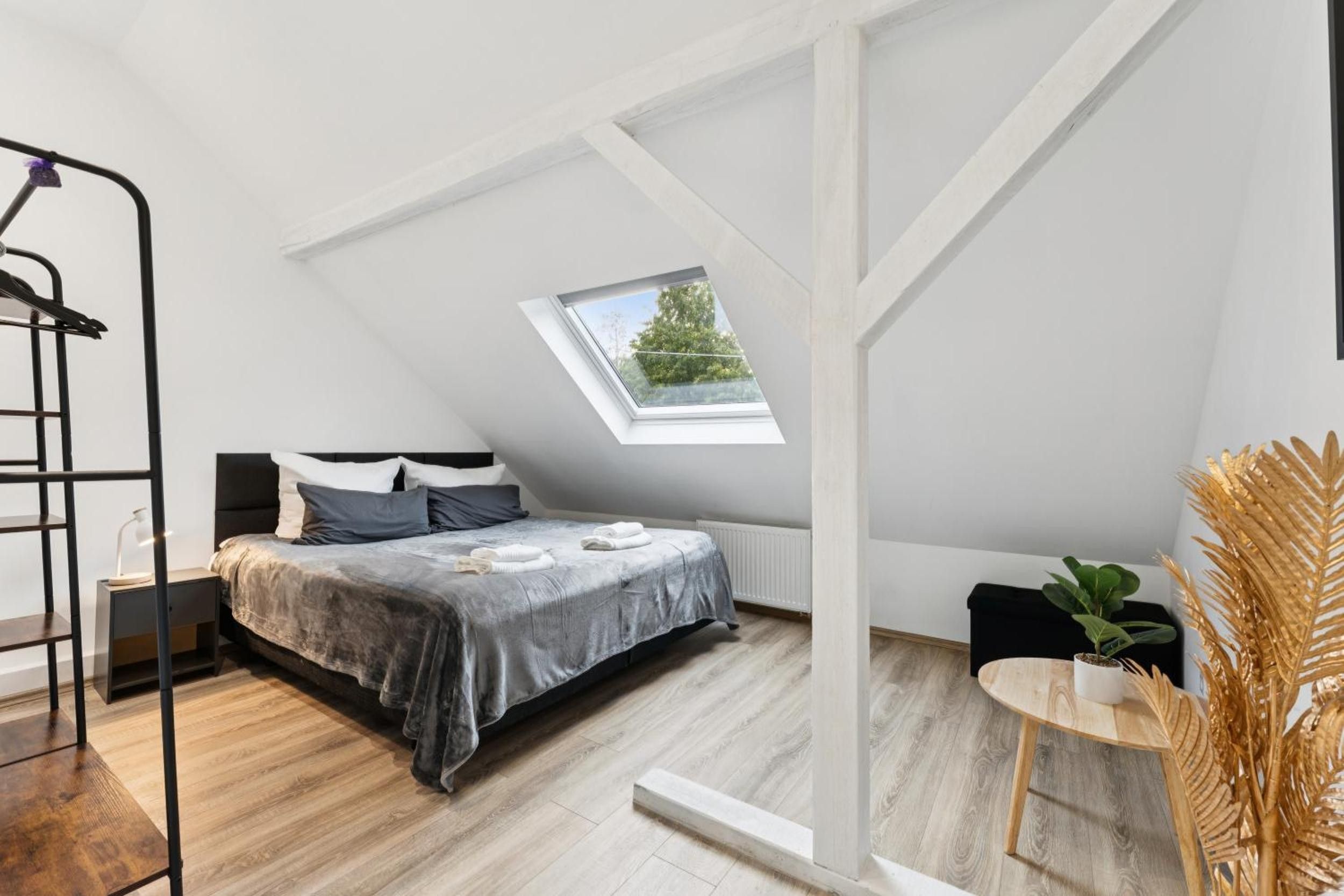 Ein Schlafzimmer mit Schrägdecke, einem Oberlicht, einem Bett mit grauer Bettdecke, einem Nachttisch mit Lampe und einem kleinen runden Tisch mit einer Pflanze.
