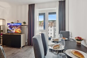 A dining area with a table set for two, a television on a stand, and a view of buildings through a window.