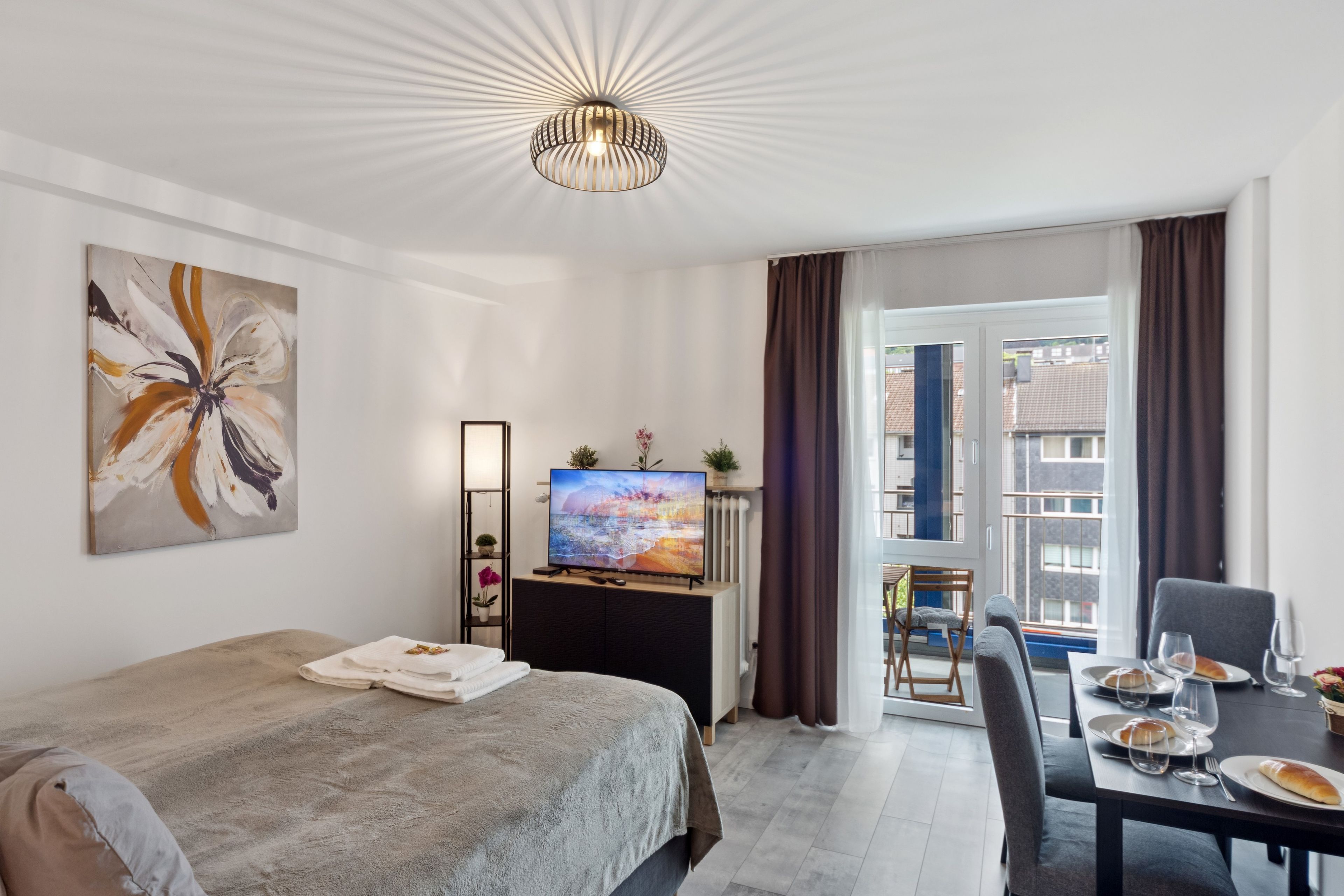A modern hotel room with a bed, a desk with a computer, a TV, and a view of the city through the window.