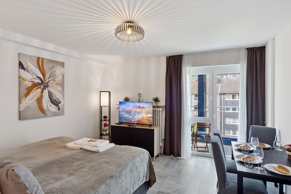 A modern hotel room with a bed, a desk with a computer, a TV, and a view of the city through the window.
