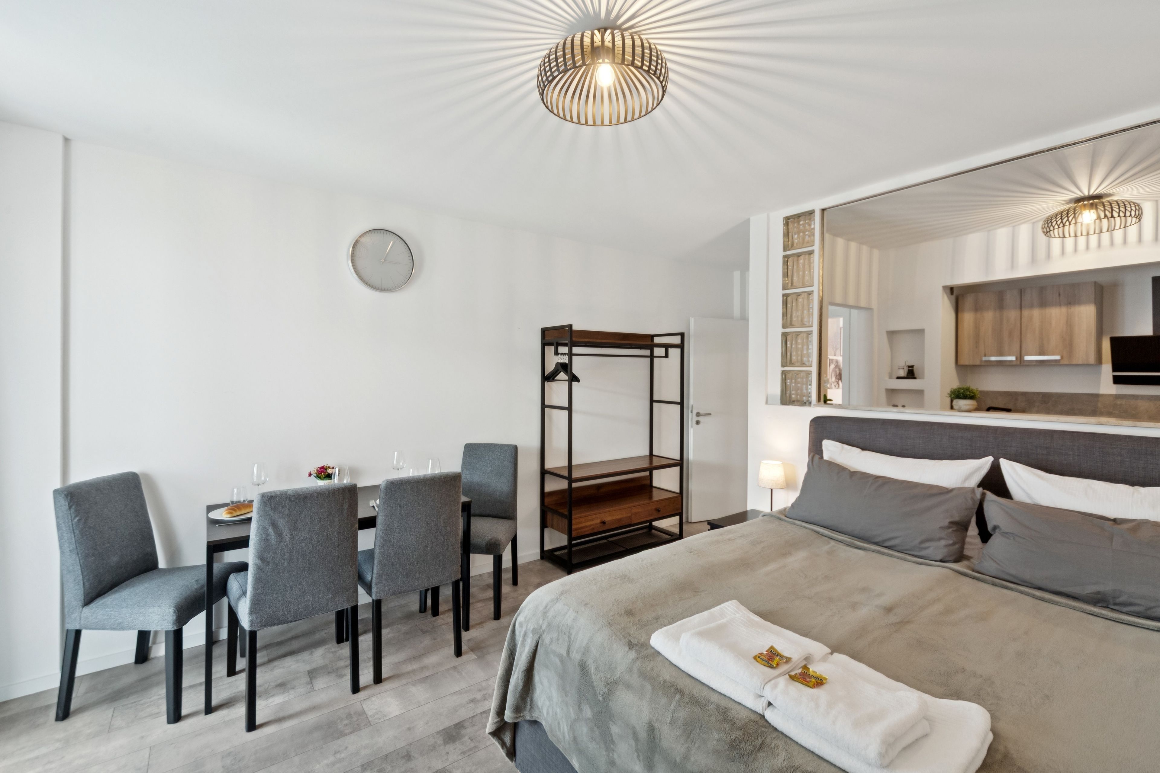 Ein modernes Hotelzimmer mit einem Bett, einem Schreibtisch und einem Sessel. An der Wand hängt ein Spiegel, darin ist eine Uhr zu sehen und im Hintergrund ein Küchenbereich.