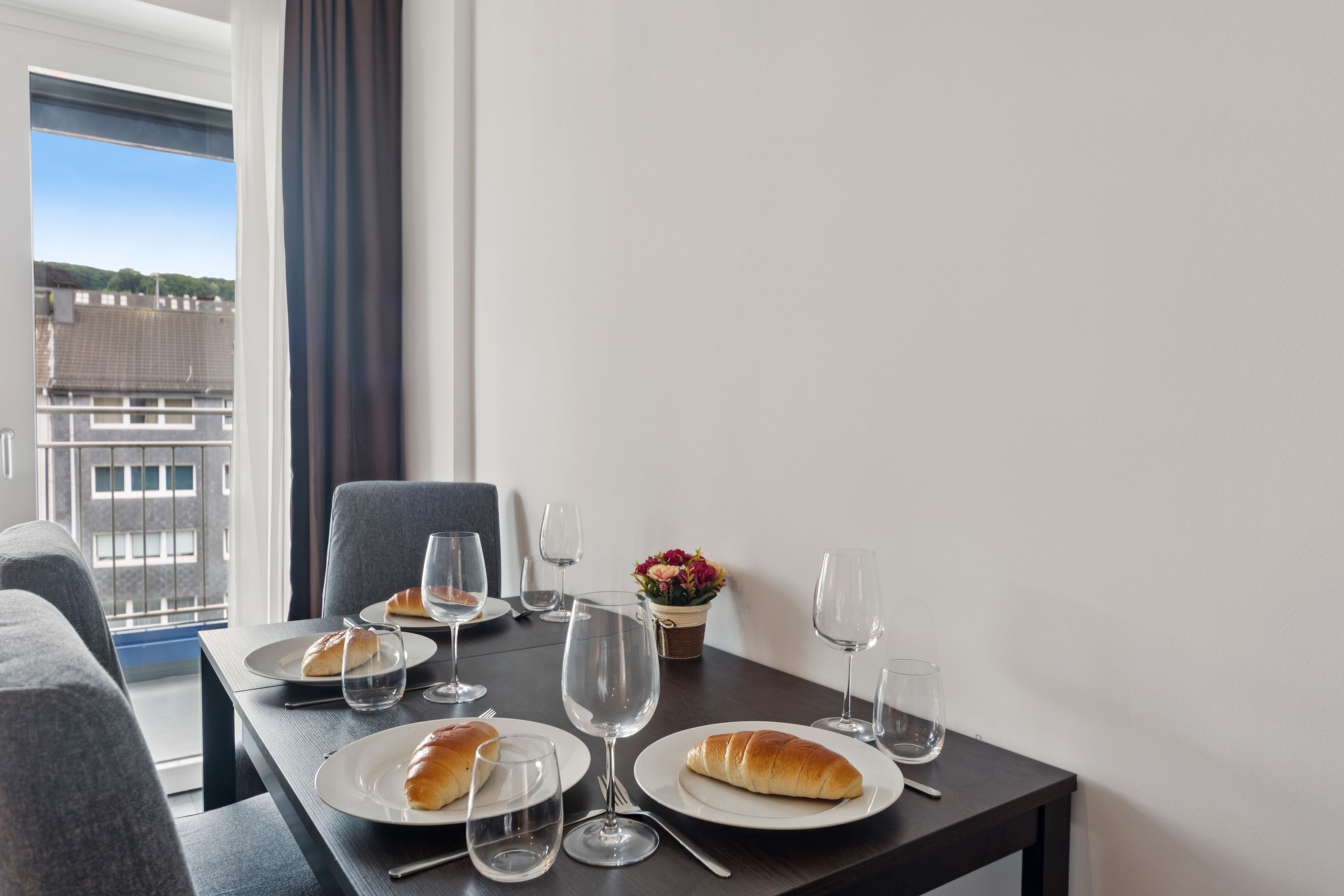 Una mesa de comedor con platos, vasos y un croissant, frente a una ventana con vistas a edificios.