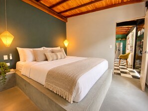 A bedroom with a large bed, bedside lamps, a plant, and a view into a kitchen area. - New Listing (Medellín)