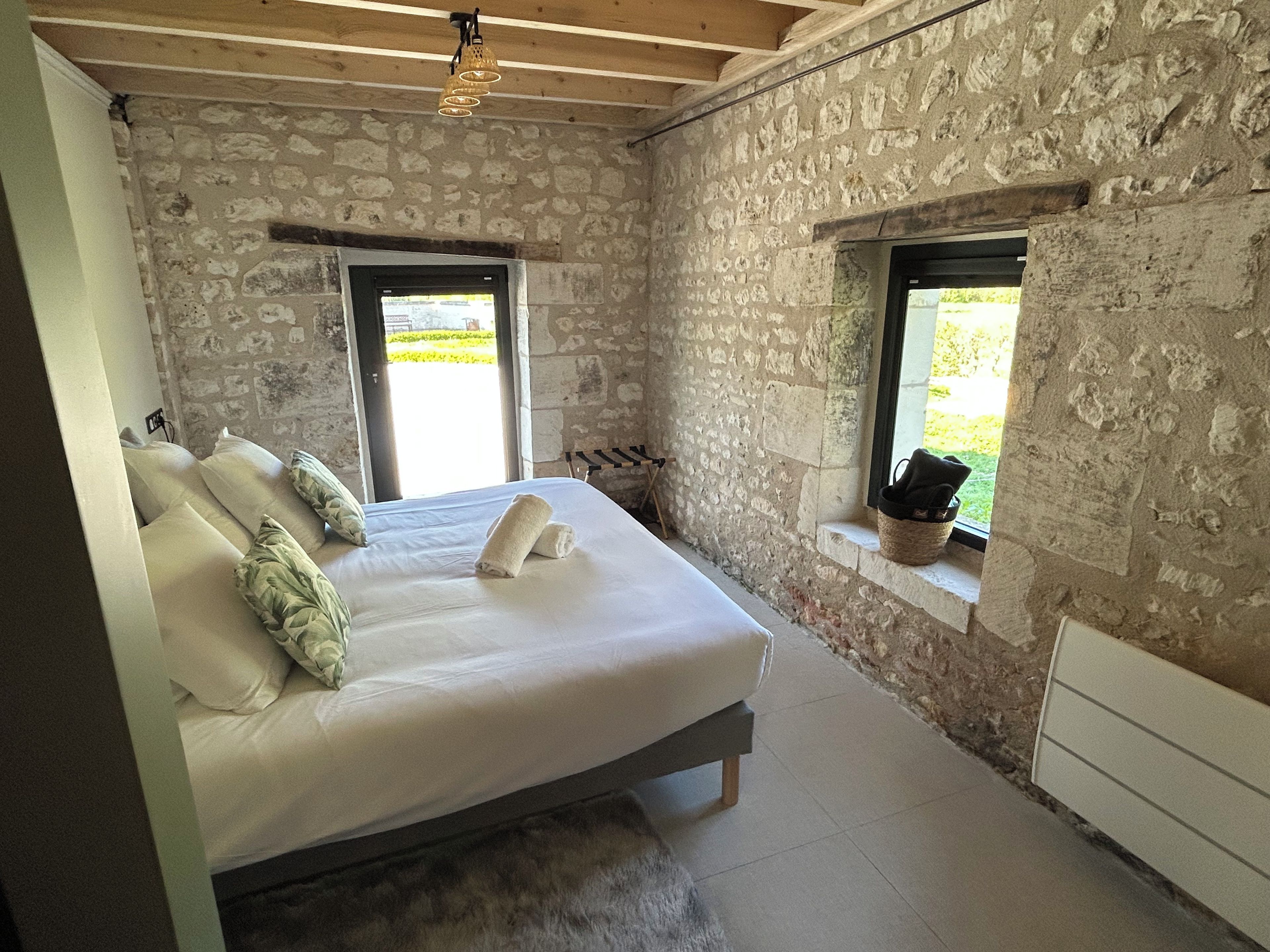A bedroom with a stone wall, a bed with pillows, a window, and a wooden ceiling.