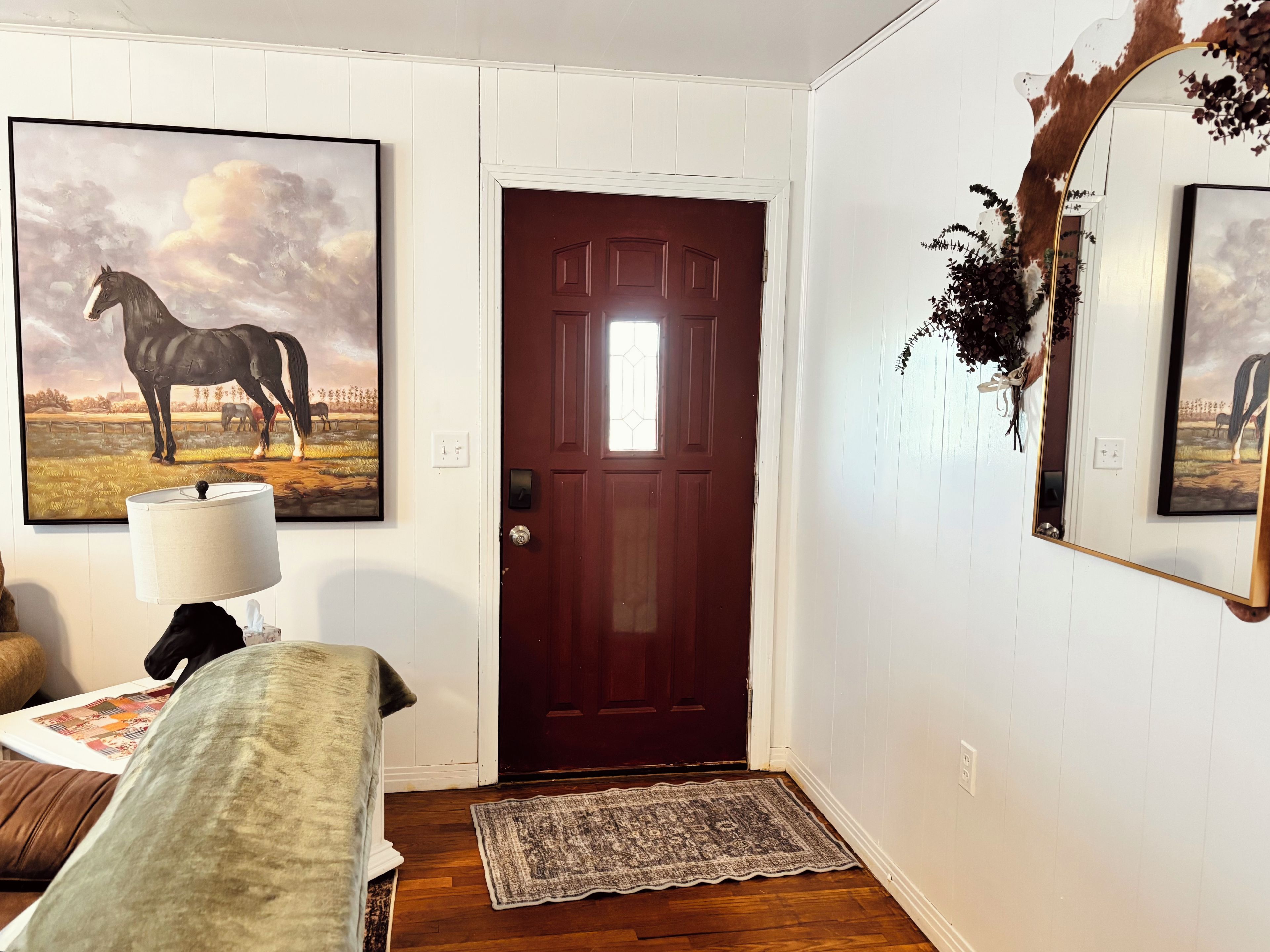 Una habitación con una puerta de madera, un cuadro enmarcado de caballos, una lámpara, un banco y una alfombra.