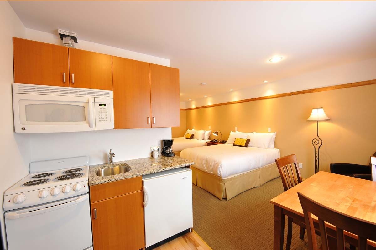 A compact hotel room with a kitchenette, a bed, a bedside table, and a lamp.