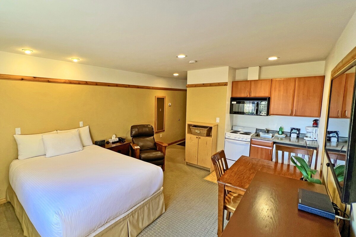 A hotel room with a bed, a desk, a chair, a kitchenette, and a bathroom.