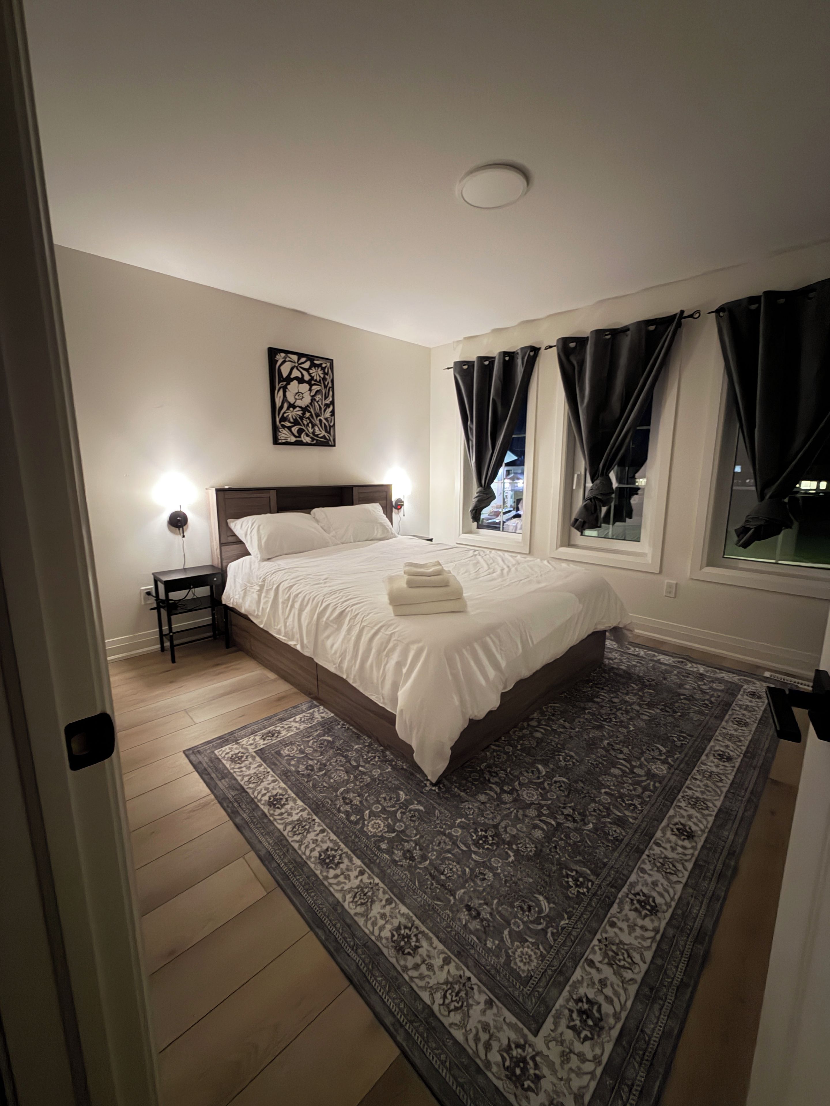 Een slaapkamer met een bed, een nachtkastje, een wandlamp, een raam met gordijnen en een kamerbreed tapijt.