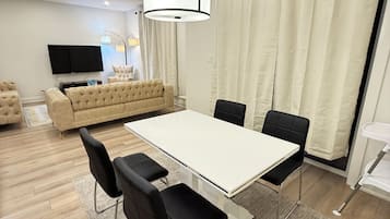 Ein modernes Wohnzimmer mit einem weißen Esstisch, schwarzen Stühlen, einem beigen Sofa und einem Flachbildfernseher.