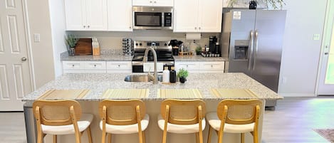A kitchen with white cabinets, a stainless steel refrigerator, a microwave, and a coffee maker. There are four wooden chairs with white cushions.