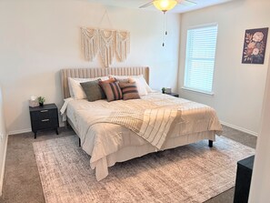 A bedroom with a bed, bedside tables, a nightstand, a hanging macrame decoration, a window with blinds, and a wall-mounted artwork. - Midland Modern 3BR Home (Midland)