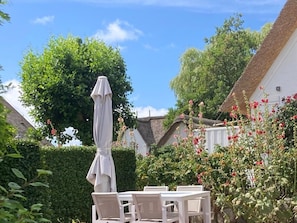 An outdoor seating area with a table and chairs, surrounded by greenery and a white building with a thatched roof. - Deichhof Holiday Apartment Luana (Dunsum)
