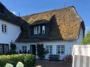 Een traditioneel huis met rieten dak, een witte gevel, een voordeur en een bank in de voortuin.