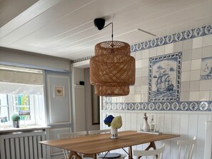Een eethoek met een houten tafel, witte stoelen, een geweven hanglamp en blauw-witte betegelde wand.