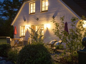 A house with a brick exterior, two windows with lights, outdoor seating, and a garden. - Aapelwin on Fohr (Oldsum)