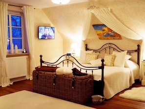 A bedroom with a four-poster bed, a wicker chest, a window with curtains, a painting on the wall, and a television on the wall. - Aapelwin on Fohr (Oldsum)