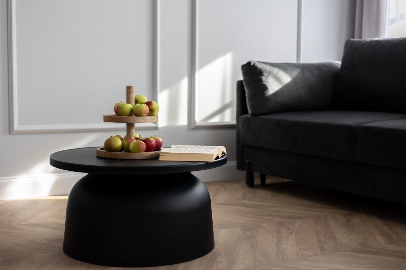 A round black table with a wooden fruit stand holding apples, a book, and a candle.