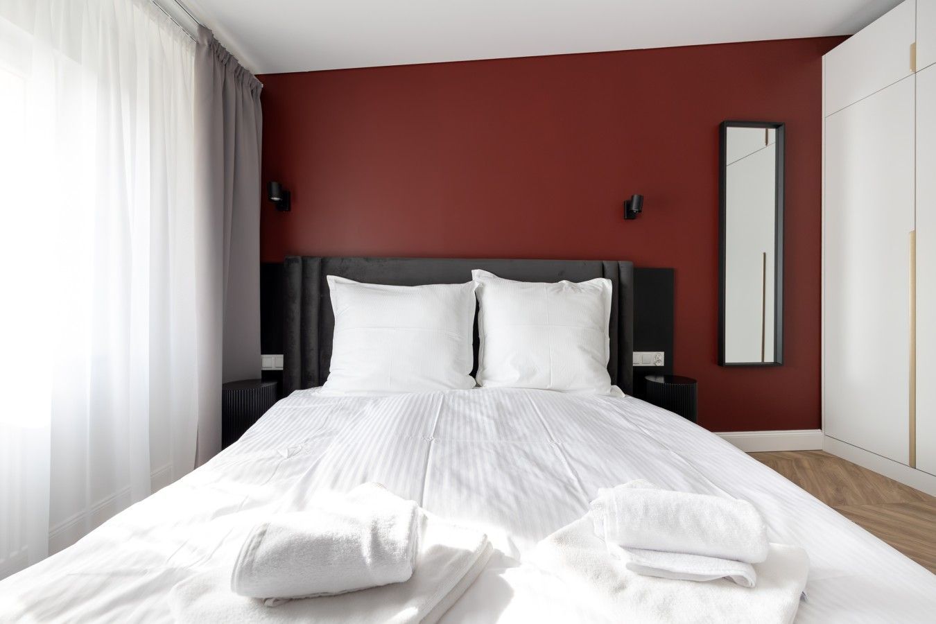 A hotel room with a bed, white linens, a dark headboard, a mirror, and a red accent wall.
