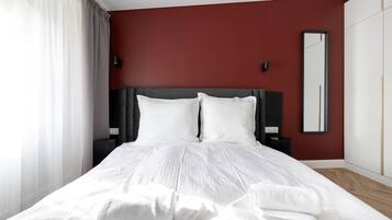 A hotel room with a bed, white linens, a dark headboard, a mirror, and a red accent wall.