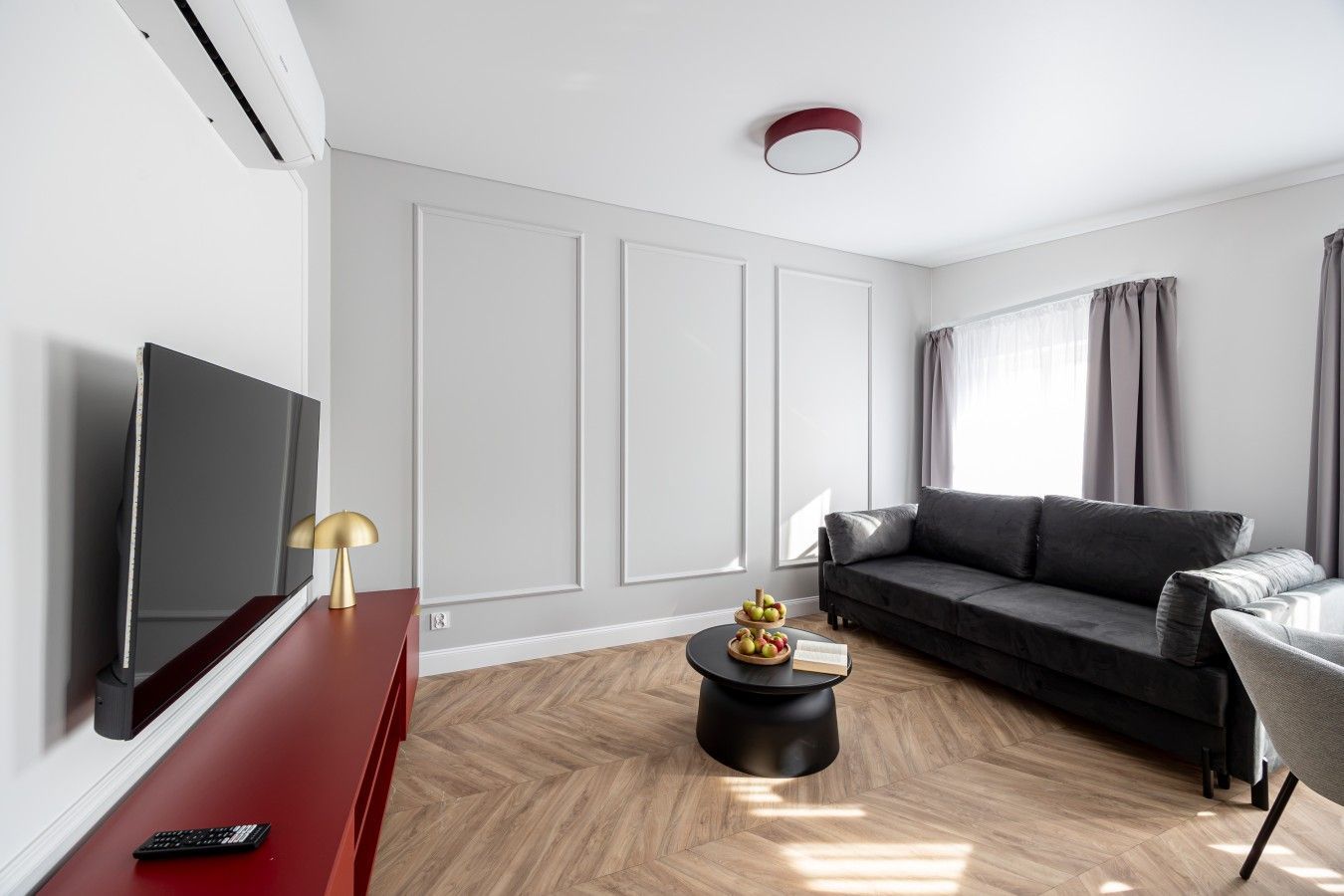 A modern living room with a flat-screen TV, a red entertainment unit, a black coffee table with fruit, a grey sofa, and a window with curtains.
