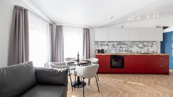 A modern living room with a grey sofa, a dining table with chairs, and a kitchen area with red cabinets and a marble backsplash.