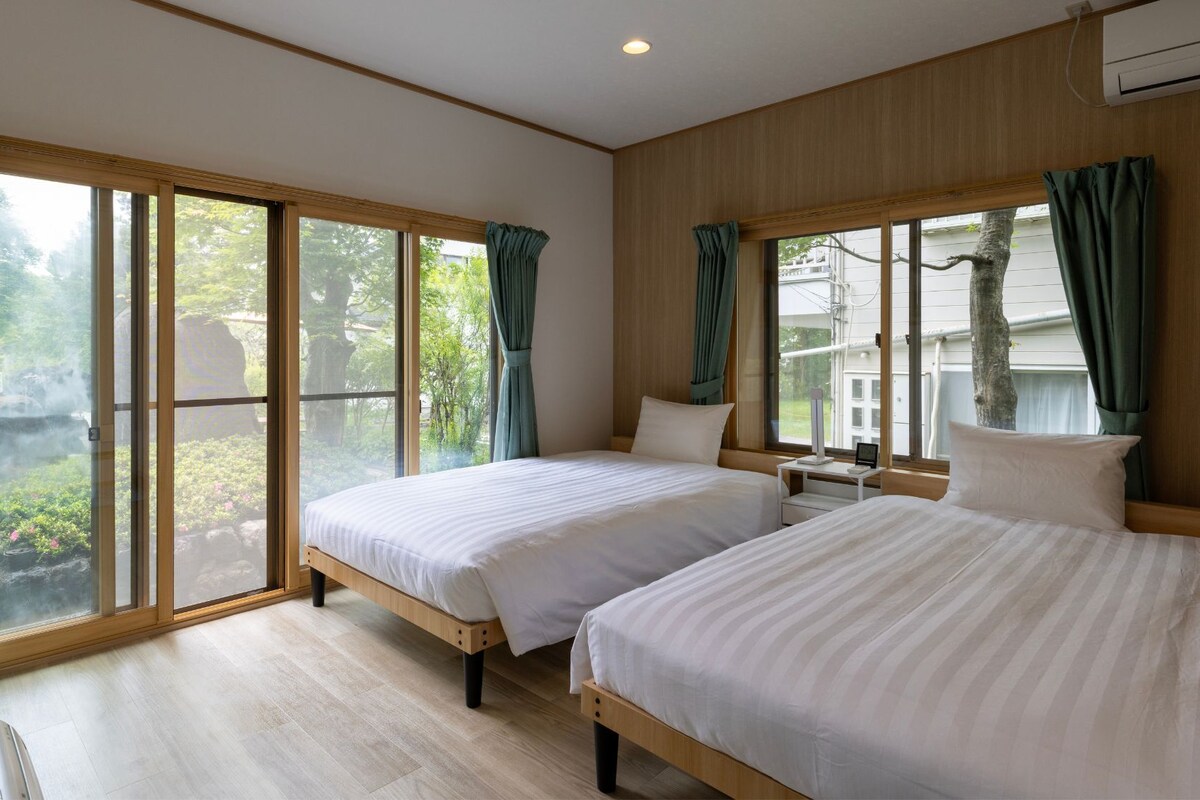 A hotel room with two beds, large windows, and a view of greenery.