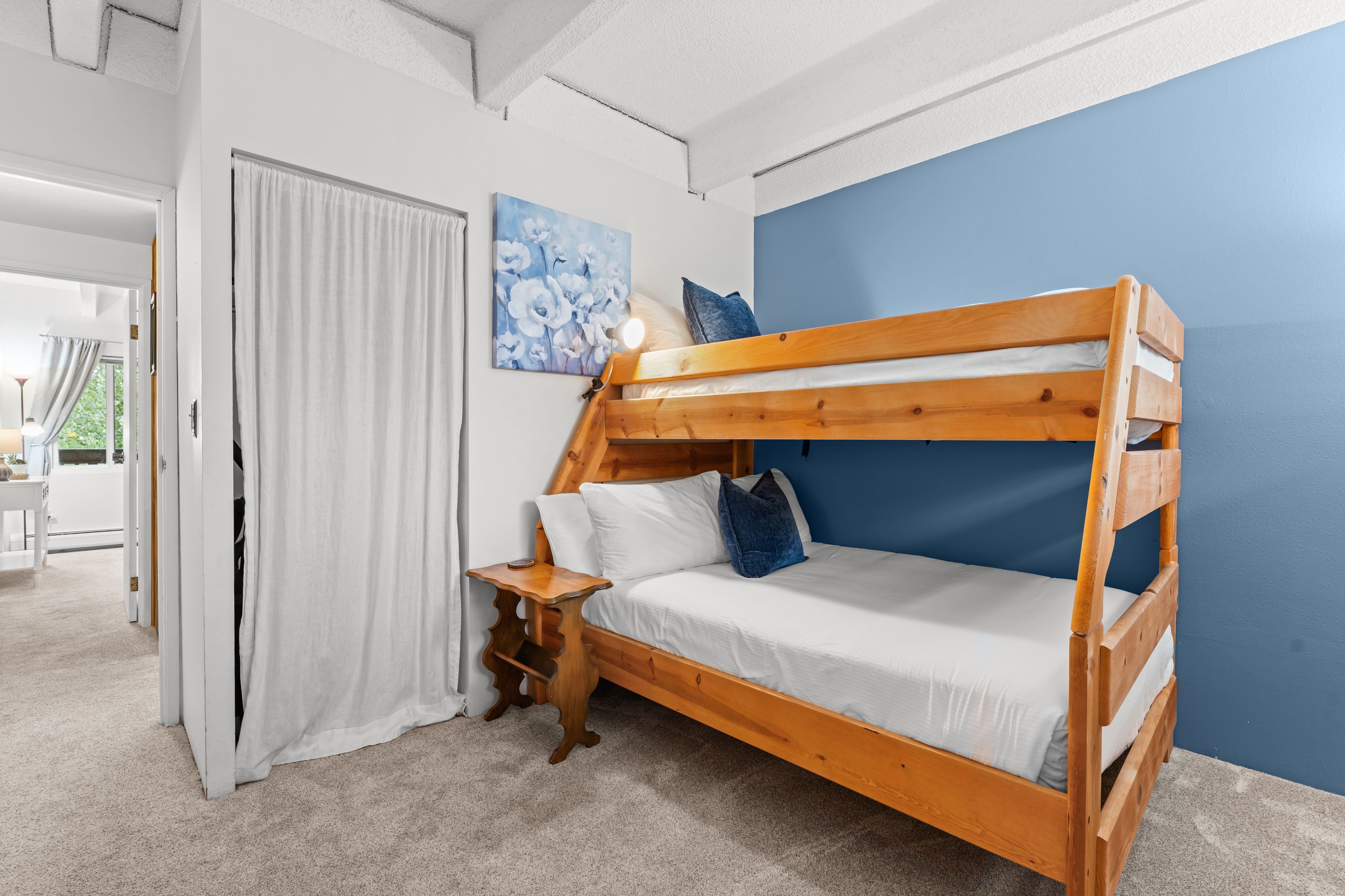 Una cama litera con estructura de madera, ropa de cama blanca y una mesita de noche con lámpara.