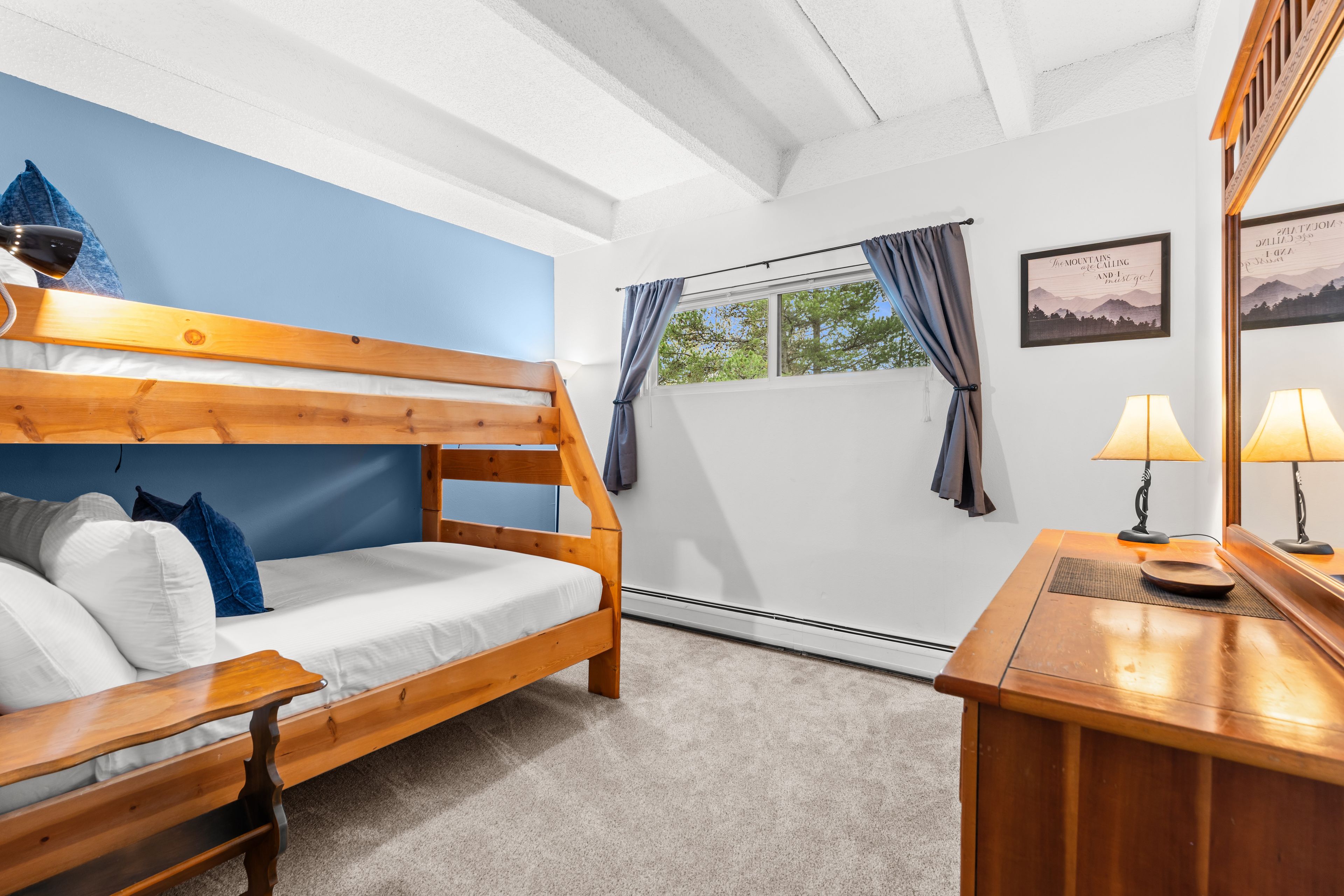 A bunk bed with a desk and lamp, a window with curtains, and a wall with a framed picture.