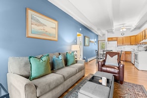 A living room with a sofa, a coffee table, and a chair. There is a kitchen in the background with wooden cabinets and a refrigerator. - Tmc304f2 (Frisco)