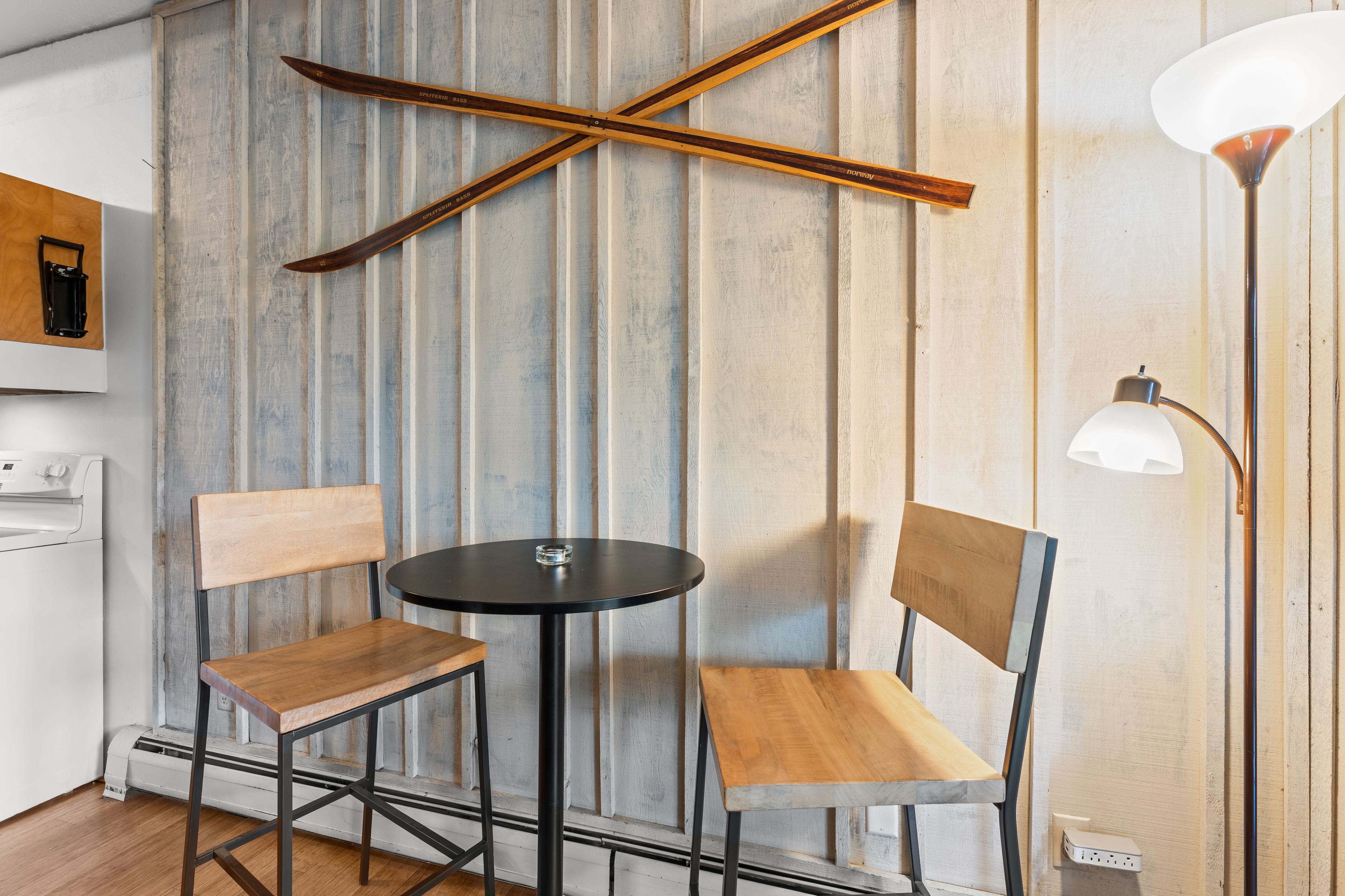 A room with a wooden-framed wall, a round table with two chairs, a floor lamp, and a washing machine.