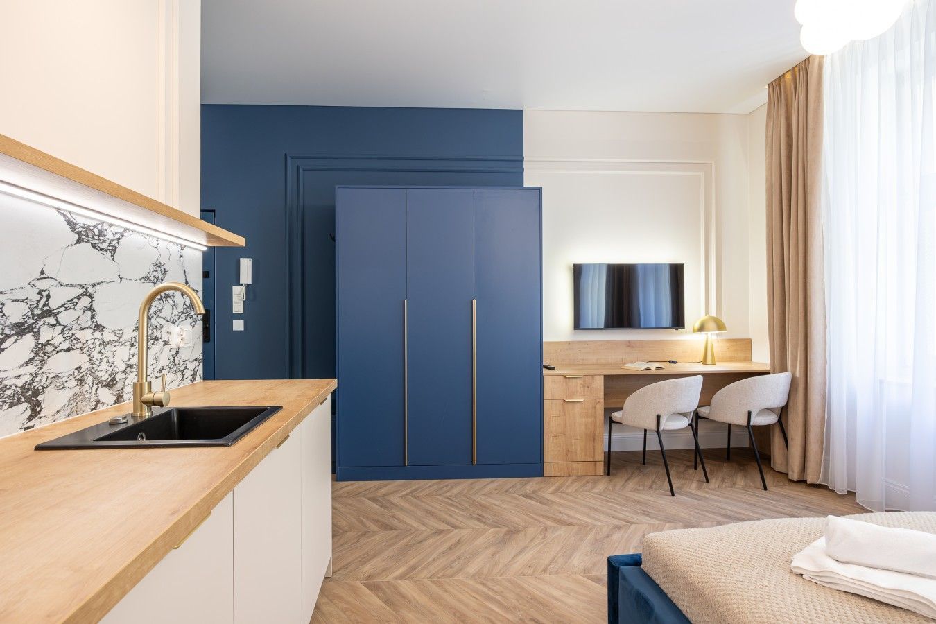 A compact hotel room with a kitchenette, a bed, a desk with a chair, and a TV.