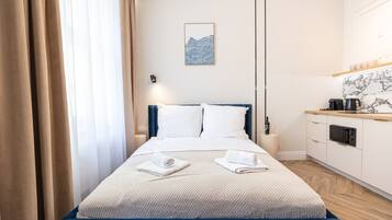 A hotel room with a bed, a bedside table, a small kitchen area, and a window with curtains.