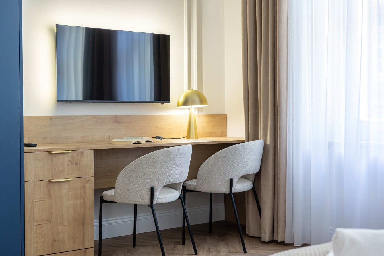 A modern hotel room with a wooden desk, two chairs, a flat-screen TV, and a lamp.