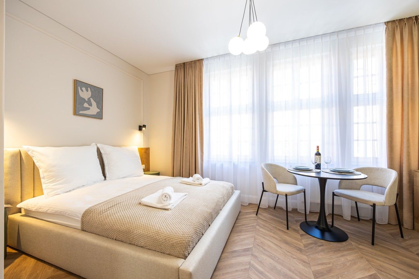 A hotel room with a large bed, a dining table with chairs, a chandelier, and a painting on the wall.