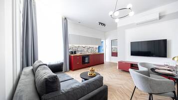 A modern living room with a sofa, a dining area with a table and chairs, a kitchen with red cabinets, and a large flat-screen TV mounted on the wall.