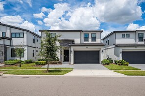 A modern residential building with a garage, a small tree in the front yard, and a clear sky. - 8BR Themed Villa Pool, Game Room & Theater (Orlando)