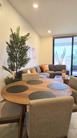 A modern living room with a round wooden table, grey upholstered chairs, a grey sofa with orange cushions, and a large green plant. - Brand New, A/C, Starlink, Terrace, 5 auto Pkg private, & Pet friendly (Ensenada)