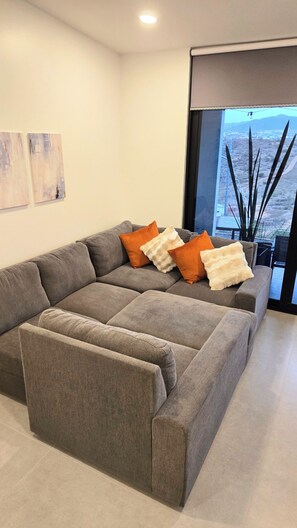 A modern living room with a grey sectional sofa, white and orange throw pillows, and a large window with a view of a desert landscape. - Brand New, A/C, Starlink, Terrace, 5 auto Pkg private, & Pet friendly (Ensenada)
