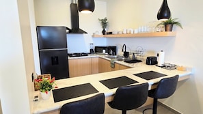 A modern kitchen with a black refrigerator, a microwave, a stove, and a sink. There are two black bar stools and a potted plant on the counter. - Brand New, A/C, Starlink, Terrace, 5 auto Pkg private, & Pet friendly (Ensenada)