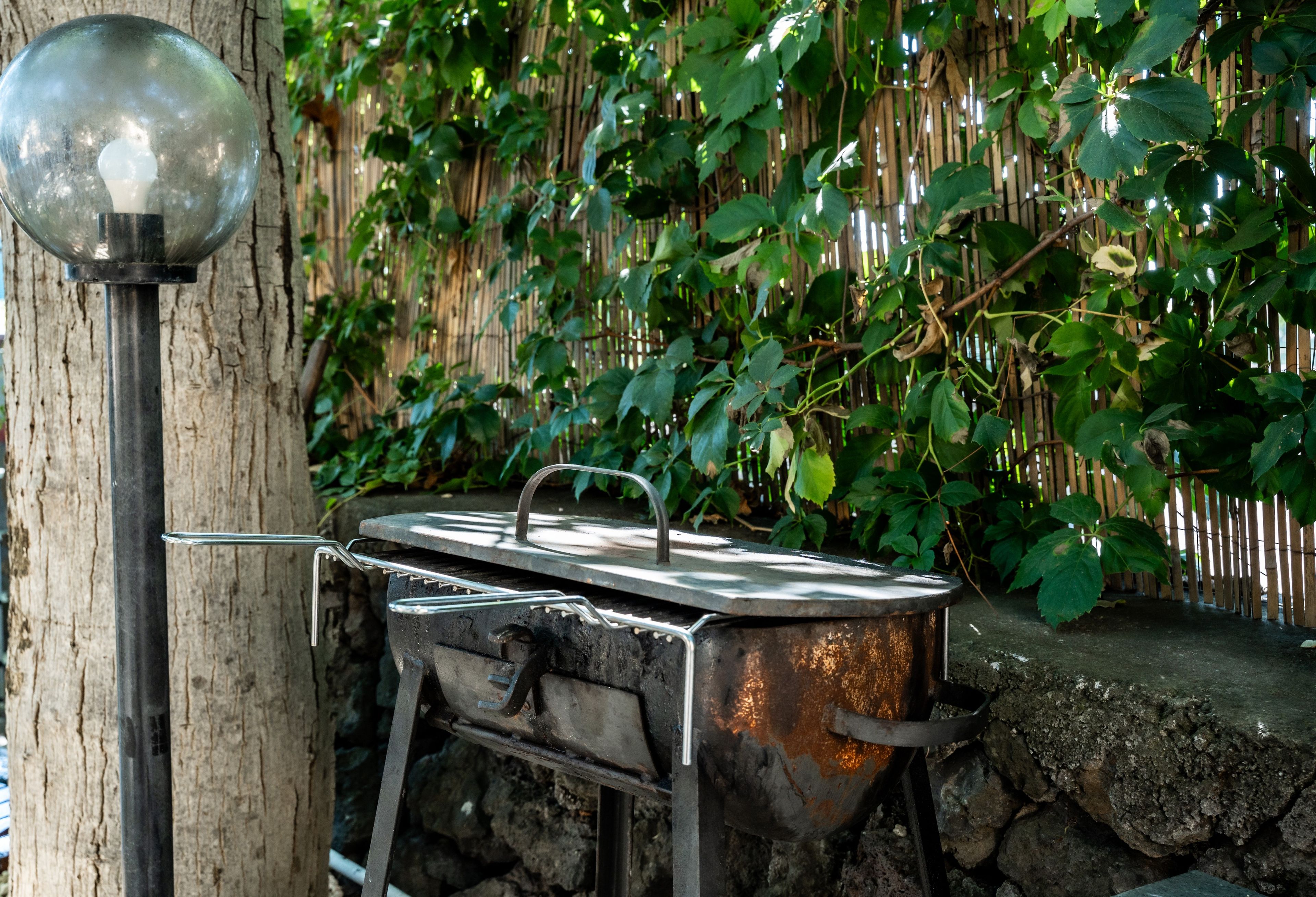 Ein Metall-Grill mit Deckel, neben einem Baum und einem Bambuszaun platziert.