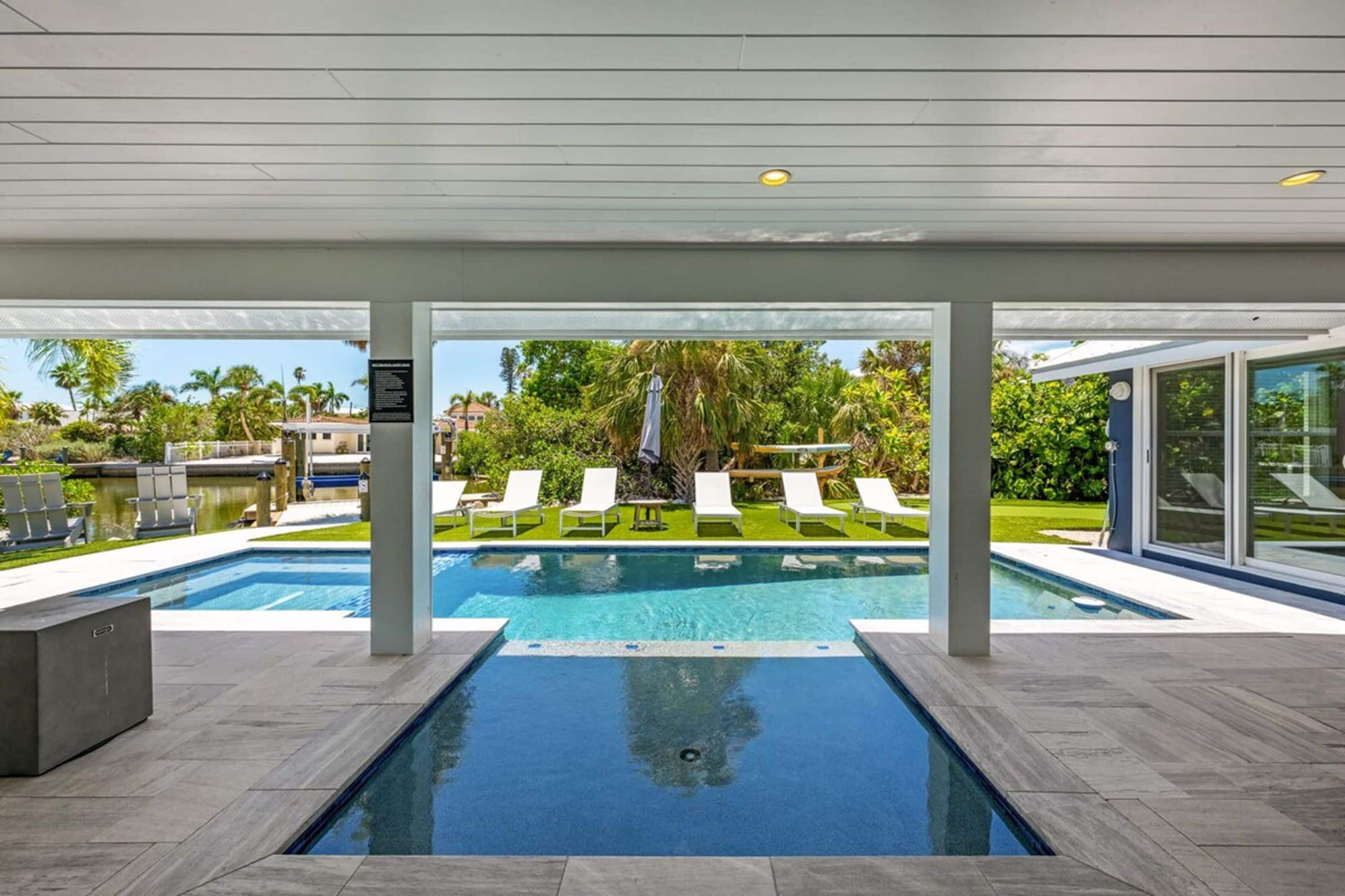 A modern indoor swimming pool with clear blue water, surrounded by lounge chairs and a view of a lush garden.