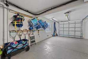 A garage with a concrete floor, a white door, and various items including a cart with a beach-themed cover, several bags, and sports rackets. - Waves - Heated Pool -steps to Beach -golf Cart - Putting Green (Anna Maria)