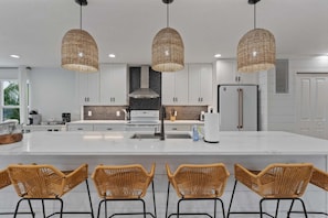 A modern kitchen with a large island, white cabinetry, and three wicker pendant lights. - Waves - Heated Pool -steps to Beach -golf Cart - Putting Green (Anna Maria)