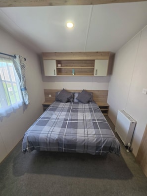 A bedroom with a bed, a wooden shelf above, a window with curtains, and a radiator.
