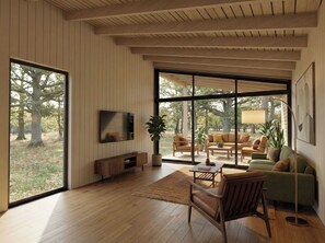 A modern living room with a large TV, wooden flooring, and a view of a garden through large windows. - Arbora 1.0 Spa (Malvern)