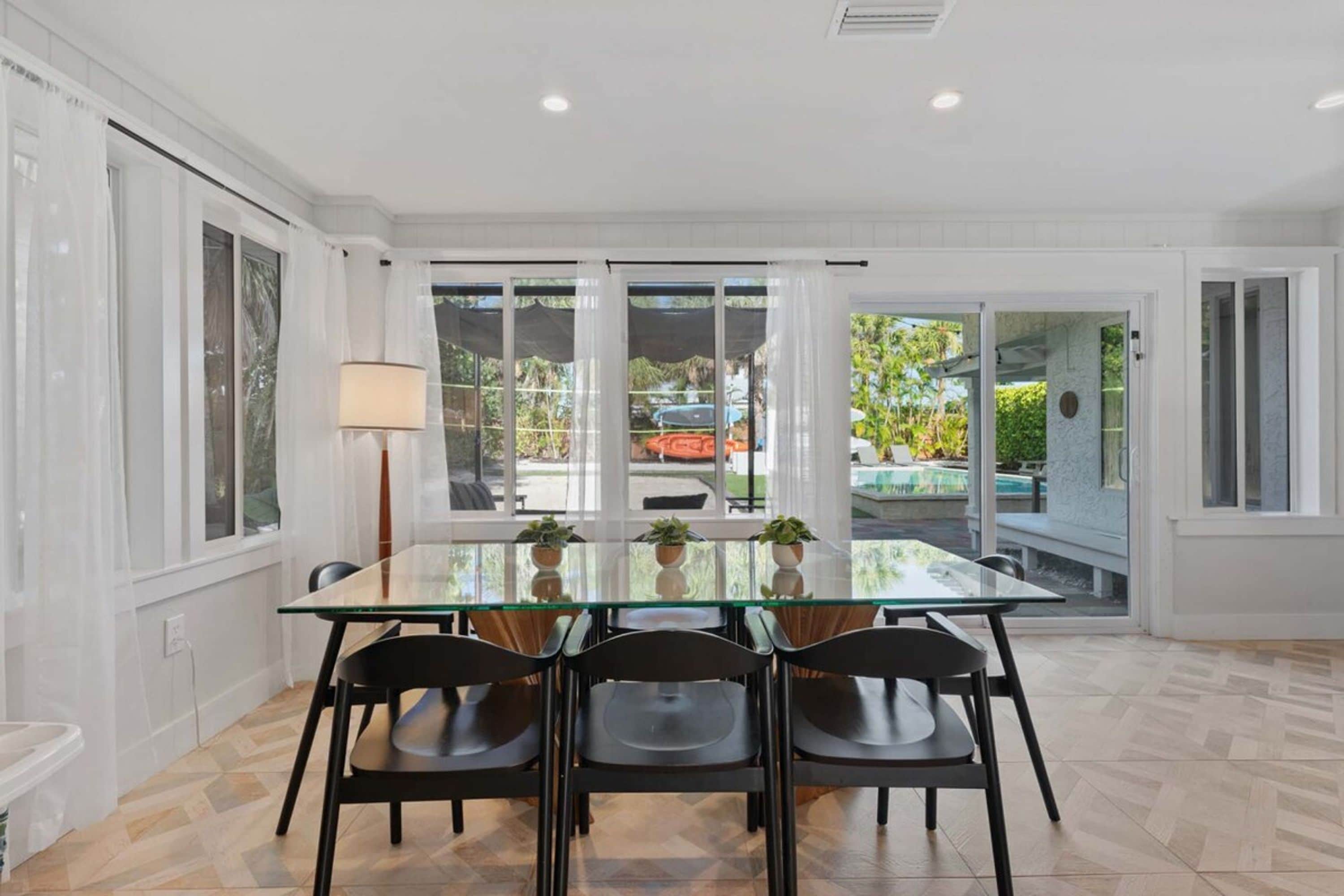 A dining area with a glass table, four chairs, and a view of a pool and garden through large sliding glass doors.