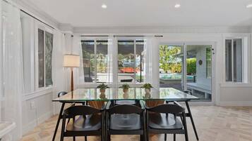 A dining area with a glass table, four chairs, and a view of a pool and garden through large sliding glass doors.