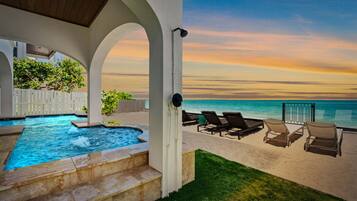 A pool area with a hot tub, lounge chairs, and a view of the ocean at sunset.