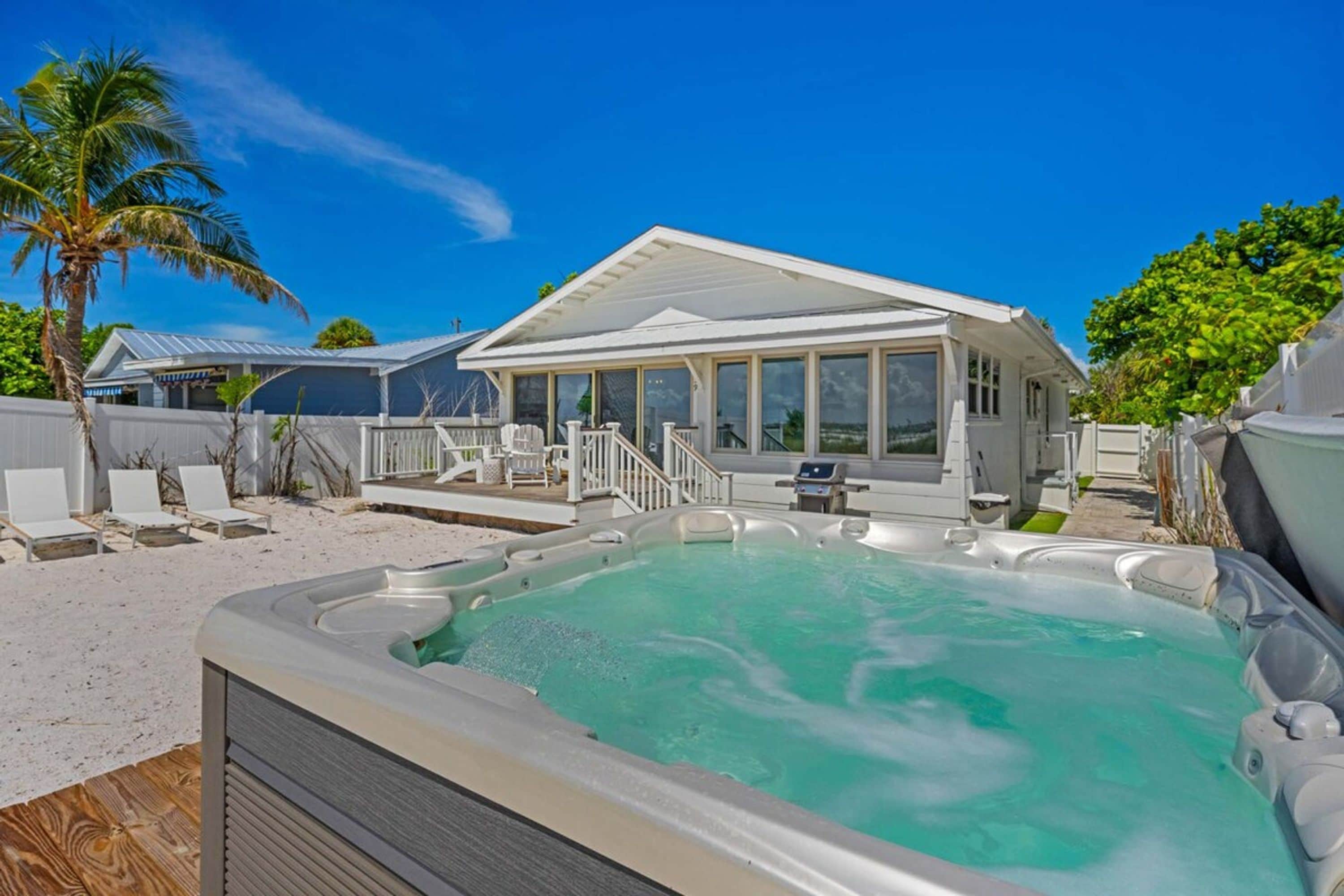 A hot tub with a deck, a covered outdoor area with chairs, and a house with a white roof.