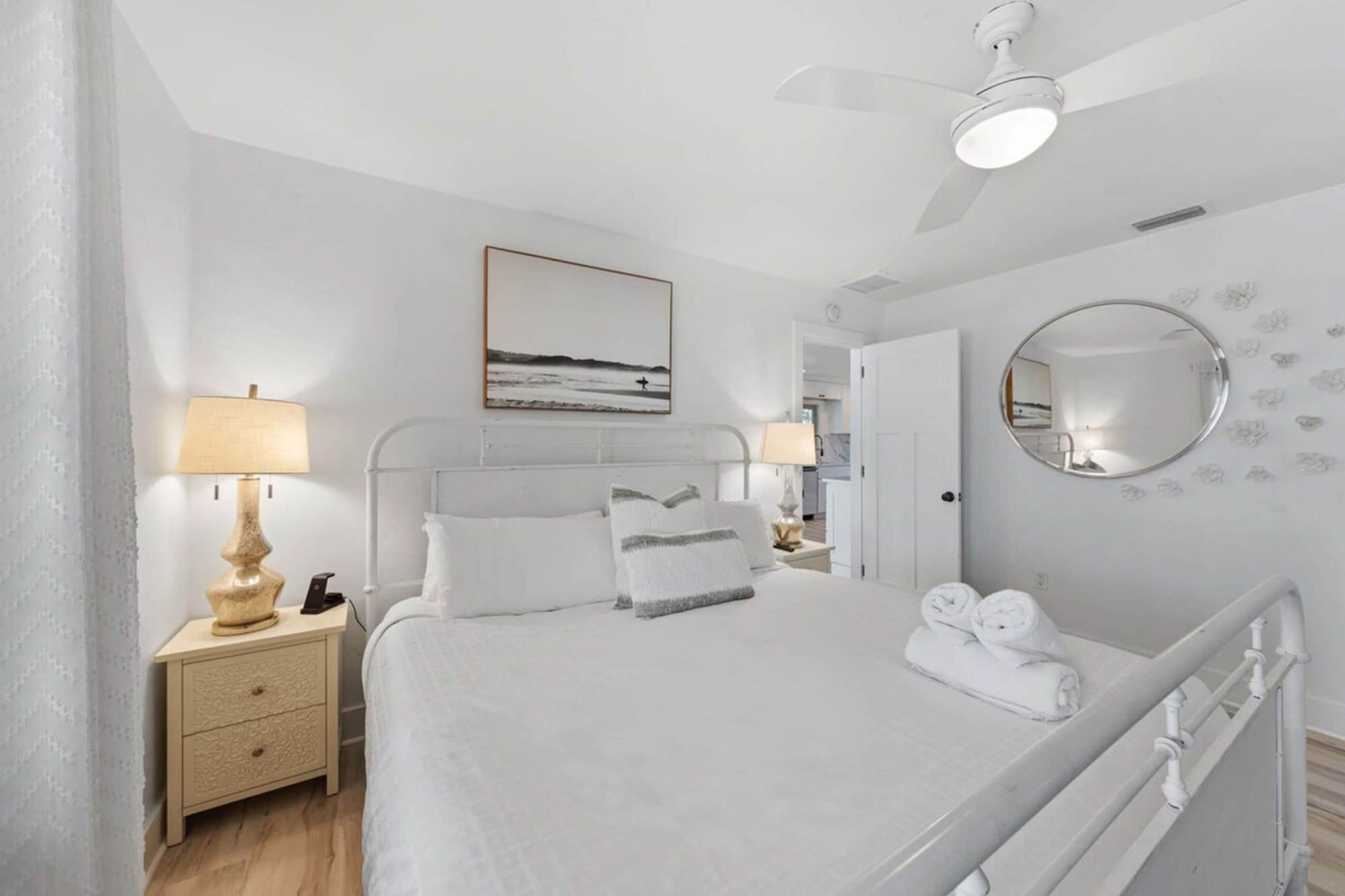 A hotel room with a bed, bedside tables, a ceiling fan, a painting, a mirror, and a view of the hallway.